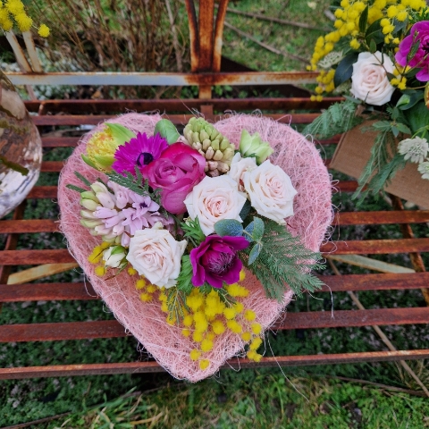 A small spring bouquet in a pink glass jam jar, edged with a pink sisal heart and filled with mimosa, tulips, anemones, roses, and hyacinths in apricot, yellow, pink, and fuchsia tones, arranged in a natural Floramay style.