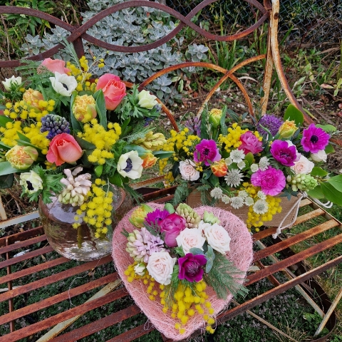 A small spring bouquet in a pink glass jam jar, edged with a pink sisal heart and filled with mimosa, tulips, anemones, roses, and hyacinths in apricot, yellow, pink, and fuchsia tones, arranged in a natural Floramay style.