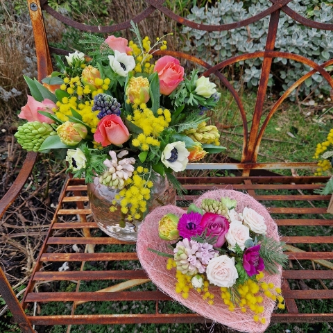 A small spring bouquet in a pink glass jam jar, edged with a pink sisal heart and filled with mimosa, tulips, anemones, roses, and hyacinths in apricot, yellow, pink, and fuchsia tones, arranged in a natural Floramay style.